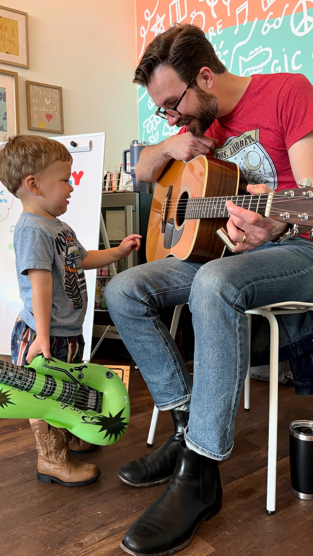 Dad playing acoustic guitar while a toddler holds an inflatable guitar