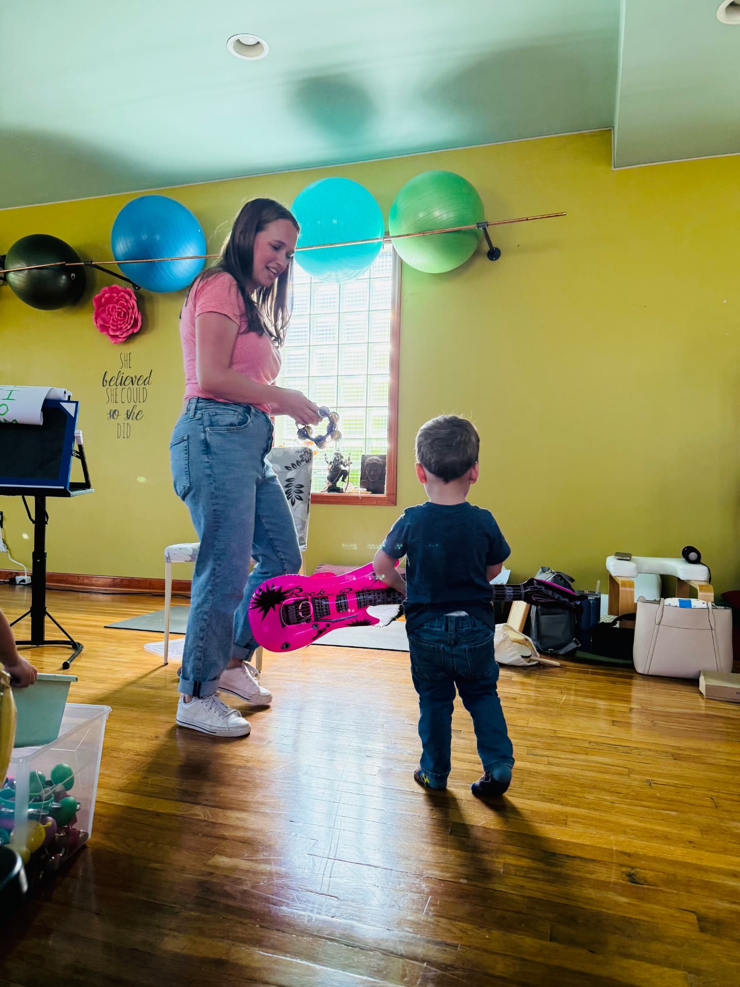 Teacher with a tambourine handing a toy guitar to a toddler in class